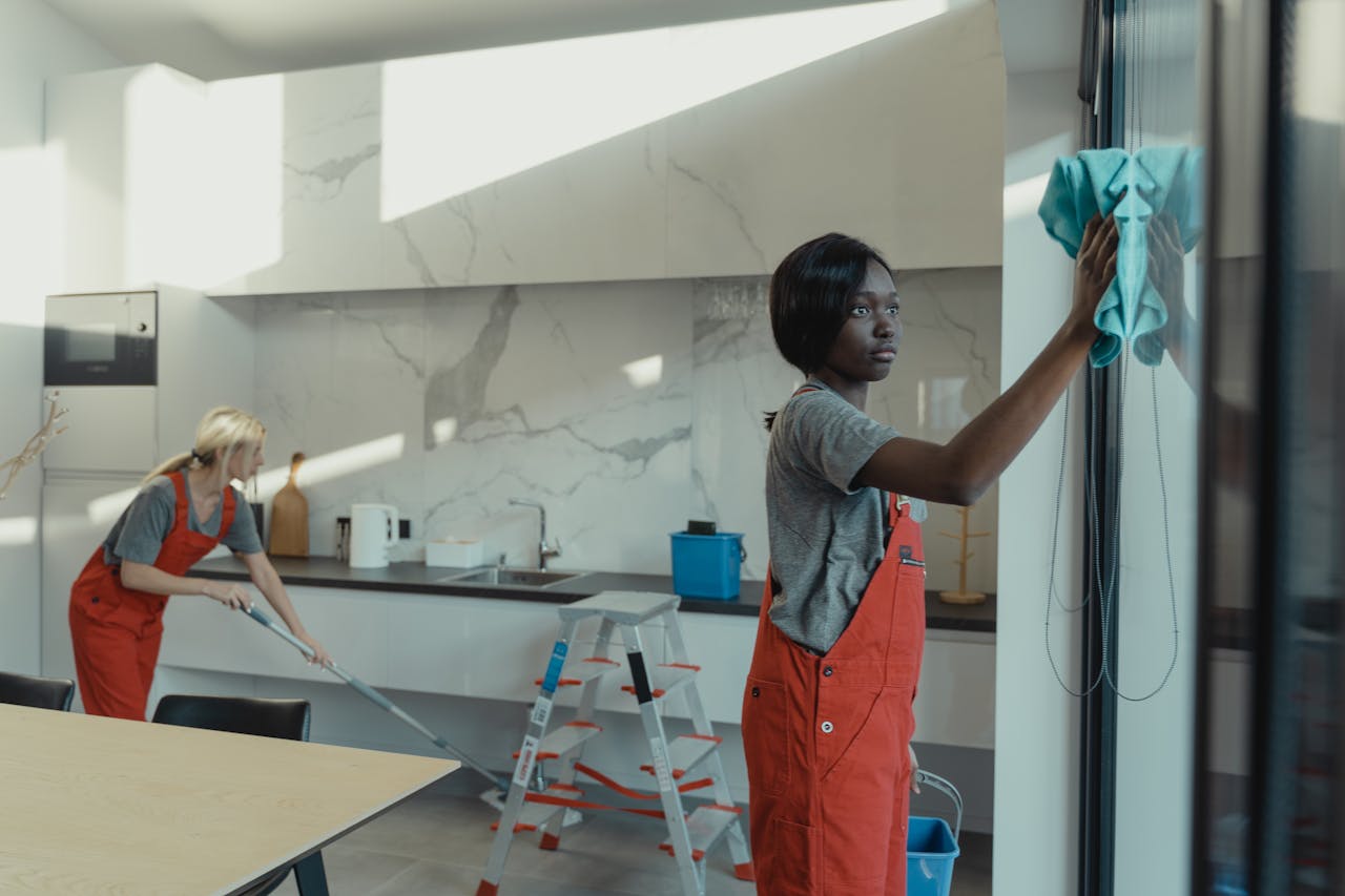 services-01 Two women cleaning a modern kitchen, wiping glass surfaces and mopping the floor.