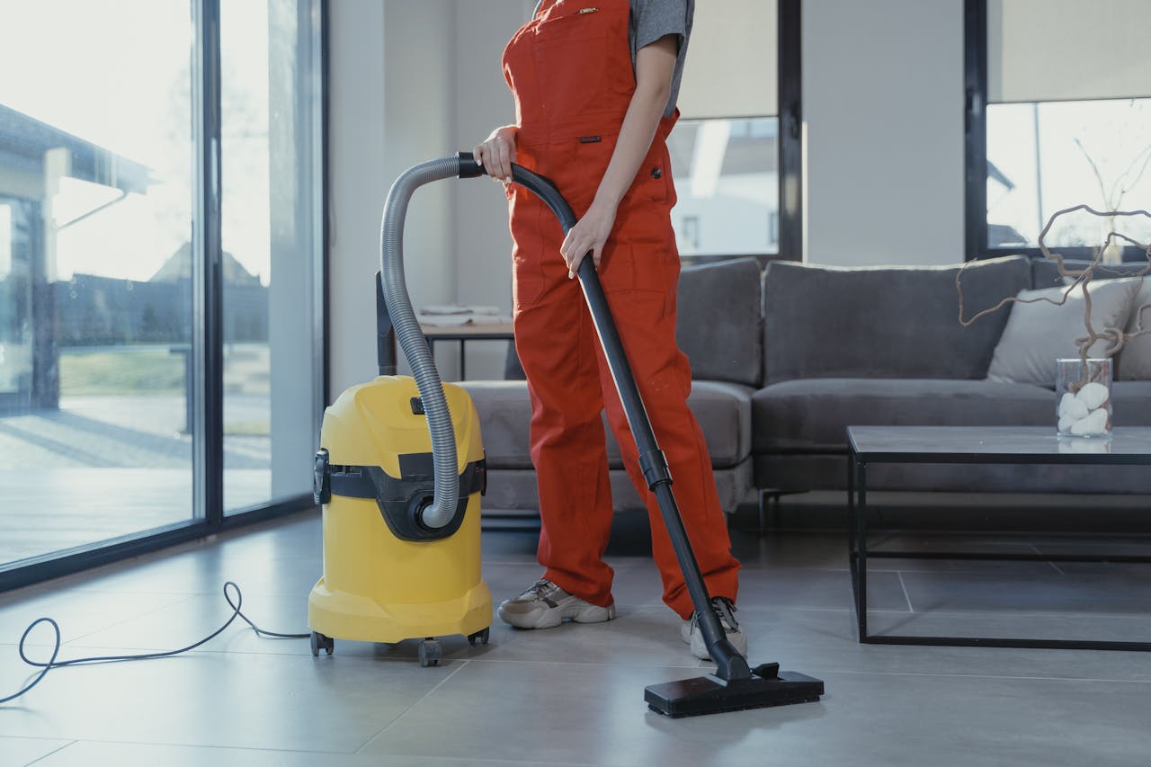 hero-img-02 A professional cleaner in red coveralls vacuuming a modern living room with a yellow vacuum cleaner.