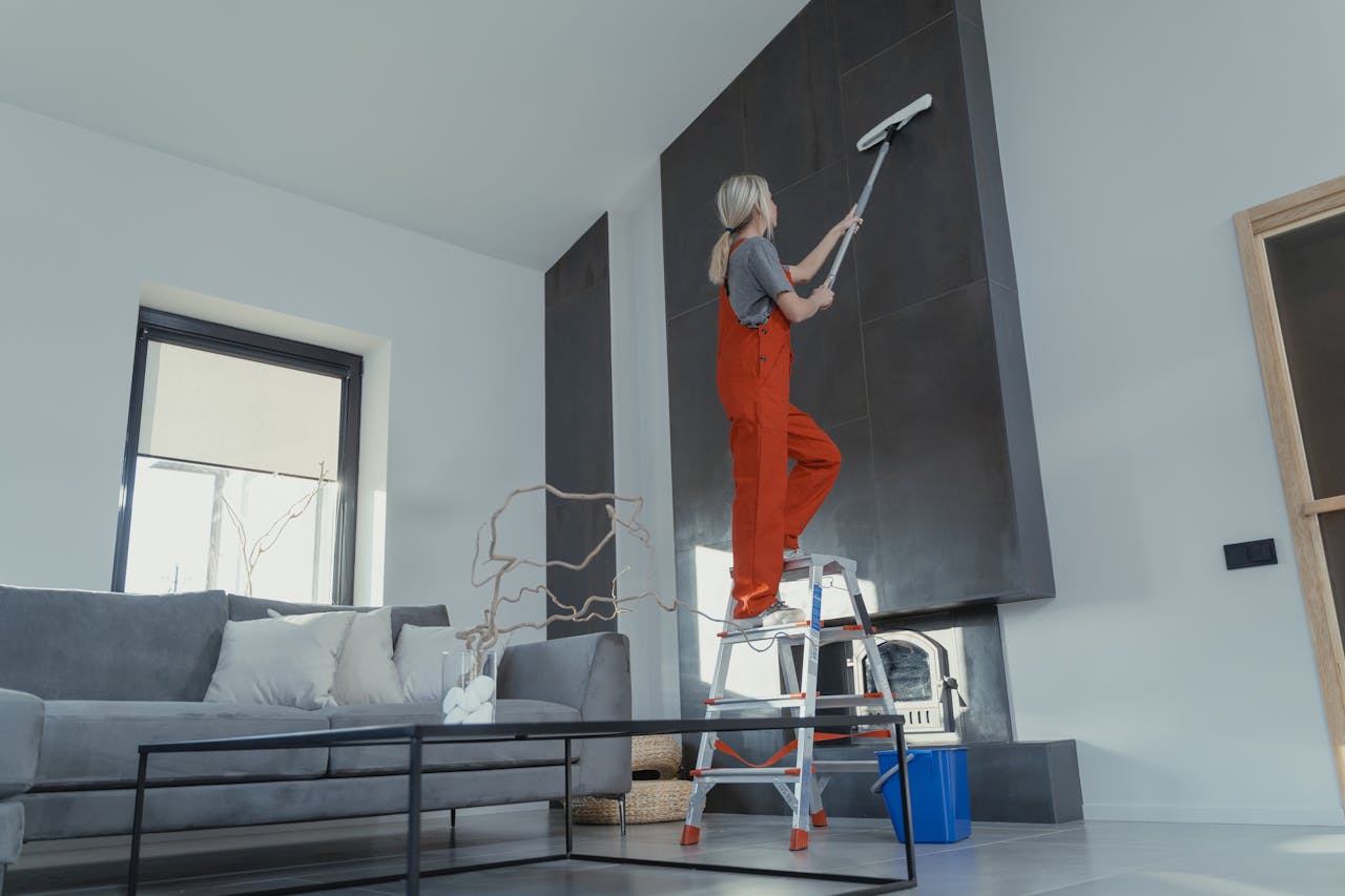 about-01 A woman stands on a ladder in a modern living room, cleaning the high wall with a mop.
